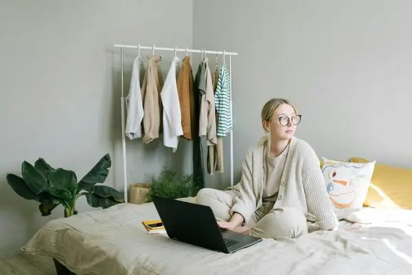 Eine Person sitzt mit einem Laptop auf einem Bett und blickt zur Seite. Im Hintergrund hängt Kleidung an einem Ständer. Auf dem Boden steht eine Pflanze.