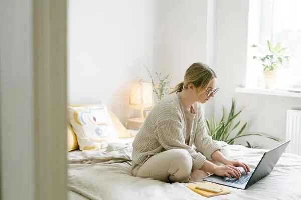 Person, die in einem hellen Raum auf einem Bett sitzt und produktiv im Homeoffice an einem Laptop arbeitet.