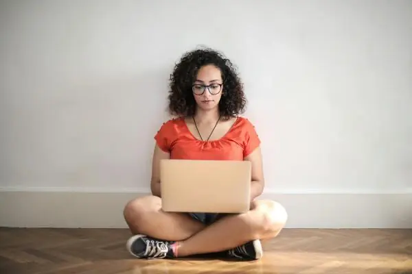 Auf dem Boden sitzende Person, trägt ein rotes Hemd und eine Brille und benutzt einen Laptop.