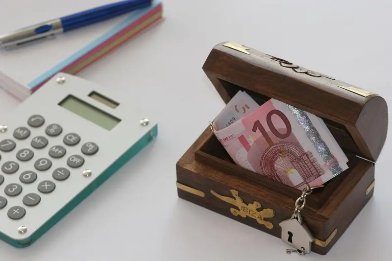 Eine kleine Holztruhe mit Euro-Banknoten steht neben einem Taschenrechner, Stiften und Notizbüchern auf einer weißen Fläche und zeigt Wege auf, wie man Abo-Kosten senken kann - vielleicht mit einem Open-Source-Tool oder Self-Hosting-Lösungen.