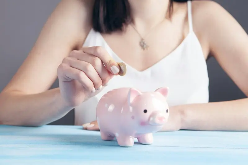 Eine Frau in einem weißen Oberteil legt eine Münze in ein rosa Sparschwein auf einem blauen Tisch und symbolisiert damit, wie Abo Kosten senken mit einem Open Source Tool Ihre Ersparnisse erhöhen kann.